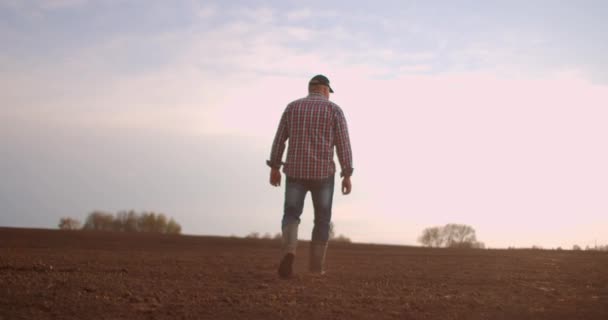En äldre manlig bonde i mössa går genom ett nyplanterat fält. Titta på den färska jorden. I slow motion gå på marken i stövlar och titta in i fjärran. — Stockvideo