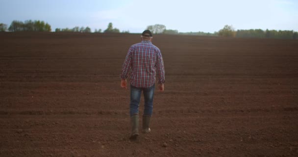 Un fermier âgé dans une casquette marche à travers un champ nouvellement planté. Regarde la terre fraîche. Au ralenti marcher sur le sol en bottes et en regardant dans la distance . — Video