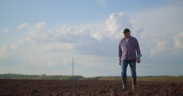 Mann Bauer geht in Gummistiefeln auf eine grüne Wiese in den Sonnenstrahlen bei Sonnenuntergang. Landwirtschaftlicher Anbau. Ein älterer Bauer geht durch ein neu gepflanztes Feld. Blicke auf die frische Erde — Stockvideo