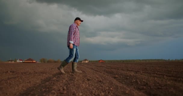 An elderly male farmer in a cap is walking through a newly planted field. Looks at the fresh earth. In slow motion walking on the ground in boots and looking into the distance. — Stock Video