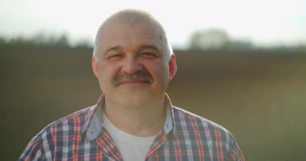 Cámara lenta: Primer plano del granjero anciano caucásico de buen aspecto y hombre sabio con un sombrero mirando al costado, girando la cara hacia la cámara y sonriendo en el campo de trigo. Retrato — Vídeos de Stock