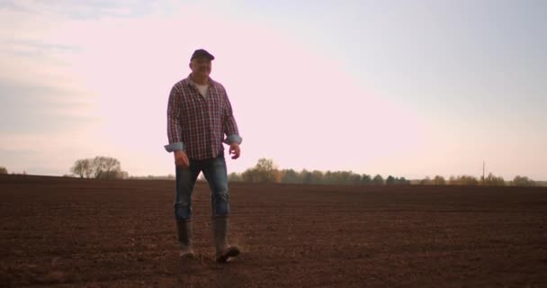 Farmer walking in idyllic,rural plowed field at sunset. Farmer wiping off sweat from forehead while walking on the field at sunset. Rubber boots for work use. A worker go with rubber boots at sunset — Stock Video