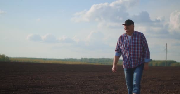 Farmer sétál idilli, vidéki szántott területen naplementekor. Farmer letörli az izzadságot a homlokáról, miközben a pályán sétál naplementekor. Gumicsizma munkahelyi használatra. Egy munkás gumicsizmában megy naplementekor. — Stock videók