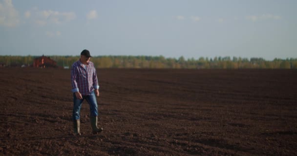 Farmář kráčí v idylickém, venkovském oraném poli při západu slunce. Farmář si při západu slunce otírá pot z čela. Gumové boty pro pracovní použití. Dělník chodí s gumovými botami při západu slunce — Stock video