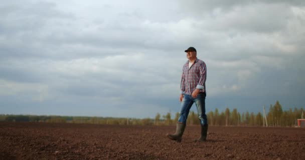 Фермер ходит в идиллическом сельском поле на закате. Фермер вытирает пот со лба во время прогулки по полю на закате. Резиновые сапоги для работы. Рабочий идет с резиновыми сапогами на закате — стоковое видео