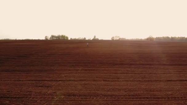 Légi felvétel arról, hogy a gazda a földjén sétál. Egy felnőtt, sapkás férfi farmer sétál át egy szántott mezőn, ültetvényekkel a csizmájában. Első látásra egy ember vizsgálja a mezőket. Gazdálkodó. — Stock videók