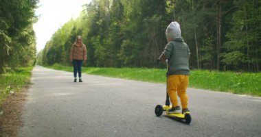 Küçük oğluna parkta scooter sürmeyi öğreten genç bir anne. Aktif aile eğlencesi. Antrenman, spor becerileri, spordaki ilk adımlar. Yavaş çekim