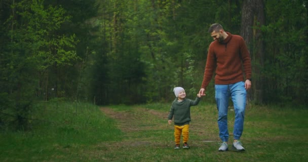 Genç bir baba ve oğul el ele tutuşarak parkta yürüyorlar. Bir adam ve bir çocuk çam ormanında birlikte yürüyorlar. Yavaş çekim mutlu baba ve oğul. Babalar Günü. 2 yaşında bir çocuk babasıyla ormanda.. — Stok video
