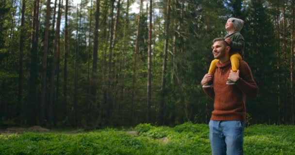 Slow motion: Son sitter på sina fäders hals i vårens soliga park. Pappadagen. Far och pojke går i skogen och son på halsen. Far och son åkte på skogsäventyr tillsammans. — Stockvideo