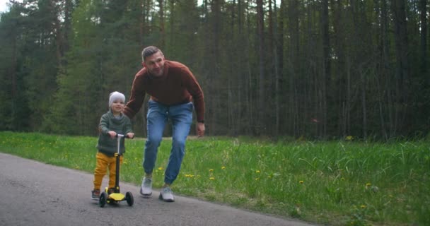スローモーションでは、幸せな若い父親が2歳の息子に、晴れた日の道路上の公園でスクーターに乗ることを教えています。公共の遊び場でスクーターに乗る愛らしい幼児の息子。父の日. — ストック動画