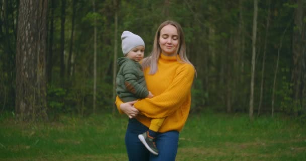 Lassított felvételen egy anya és fia sétálnak át a parkon pulóverben. Egy szerető anya a fiával a karjaiban sétál egy fenyőerdőben. Anyák napja. Kaukázusi nő egy vicces fiú 2 éves. — Stock videók