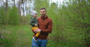Genç bir baba ve oğul el ele tutuşarak parkta yürüyorlar. Bir adam ve bir çocuk çam ormanında birlikte yürüyorlar. Yavaş çekim mutlu baba ve oğul. Babalar Günü. 2 yaşında bir çocuk babasıyla ormanda..