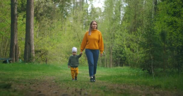 In Zeitlupe laufen Mutter und Sohn in Pullovern durch den Park. Eine liebevolle Mutter geht mit ihrem Sohn im Arm durch einen Kiefernwald. Muttertag. Kaukasische Frau mit einem lustigen Jungen von 2 Jahren. — Stockvideo