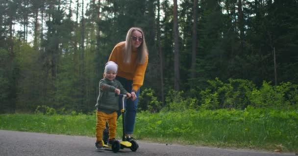 Egy fiatal anya és a fia megtanulnak robogózni. Anyák napja. Egy fenyőerdőben az úton boldog két ember egy nő és egy kisfiú együtt egy robogón. A gyermek segítése és oktatása. — Stock videók