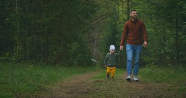 Genç bir baba ve oğul el ele tutuşarak parkta yürüyorlar. Bir adam ve bir çocuk çam ormanında birlikte yürüyorlar. Yavaş çekim mutlu baba ve oğul. Babalar Günü. 2 yaşında bir çocuk babasıyla ormanda..