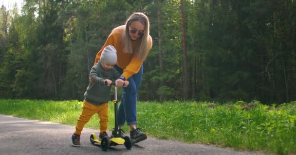 公園でスクーターに乗る方法彼女の幼児の息子を示す若い母親。アクティブな家族のレジャー。トレーニング、スポーツスキル、スポーツの最初のステップ。スローモーション — ストック動画