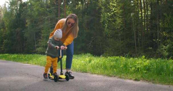 Une jeune mère et son fils apprennent à faire du scooter. La fête des mères. Dans une pinède sur la route heureux deux personnes une femme et un petit garçon ensemble sur un scooter. Aide et éducation de l'enfant . — Video