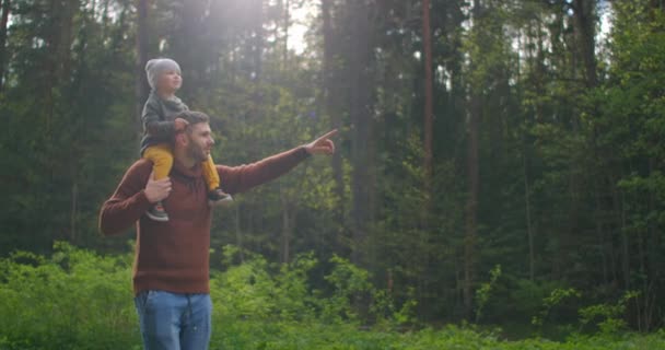 Αργή κίνηση: Πατέρας και γιος περπατούν στο πάρκο το ηλιοβασίλεμα. Ένα δίχρονο παιδί κάθεται στους ώμους των γονιών. Χαρούμενη οικογενειακή ιδέα. Πατέρας κουβαλάει γιο στους ώμους του. — Αρχείο Βίντεο