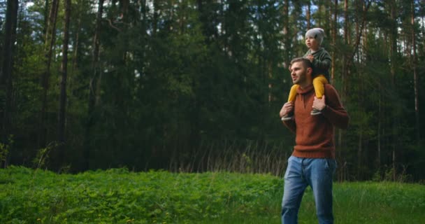 Zpomalený film: otec a syn se procházejí v parku při západu slunce. Dva roky starý kluk sedí na ramenou rodičů. Šťastný rodinný koncept. Otec nosí syna na ramenou. — Stock video