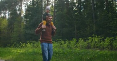 Gün batımında parkta yürüyen baba ve oğul. İki yaşında bir çocuk ebeveyn omuzlarında oturuyor. Mutlu aile kavramı. Baba oğlunu omuzlarında taşıyor..
