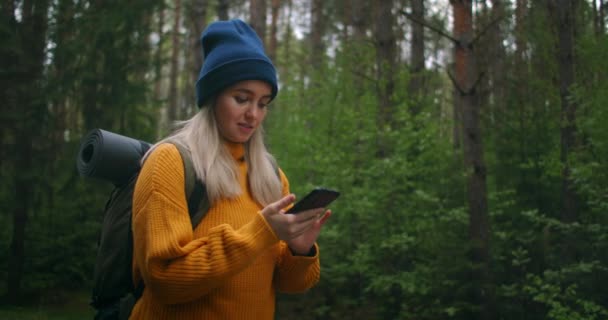En kvinnlig resenär som håller i en smartphone i en gul tröja med ryggsäck går längs en stig i skogen och tittar på vackra vyer. Fotografera med appen och Internet i skogen. — Stockvideo