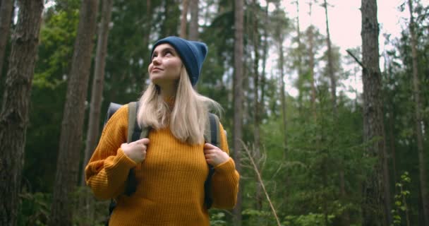 Una viaggiatrice in un maglione giallo con uno zaino sta camminando lungo un sentiero nella foresta guardando una splendida vista. Viaggiatore femminile che esplora la foresta in un maglione con uno zaino — Video Stock
