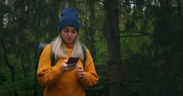 Una mujer viajera sosteniendo un teléfono inteligente en un suéter amarillo con una mochila está caminando a lo largo de un camino en el bosque mirando hermosas vistas. Tomar fotos utilizar la aplicación y el Internet en el bosque . — Vídeos de Stock