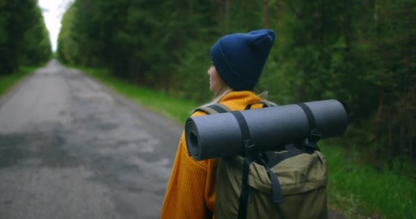 Une voyageuse dans un pull jaune avec un sac à dos marche le long d'un chemin dans la forêt en regardant de belles vues. Voyageuse explorant la forêt dans un pull avec un sac à dos — Video