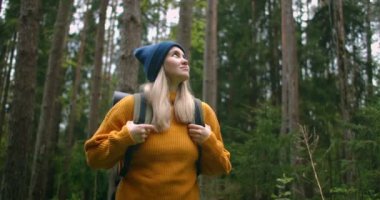 Sırt çantalı sarı kazaklı bir kadın gezgin ormanda bir patika boyunca yürüyor ve güzel manzaralara bakıyor. Sırt çantasıyla ormanı gezen bir kadın gezgin.