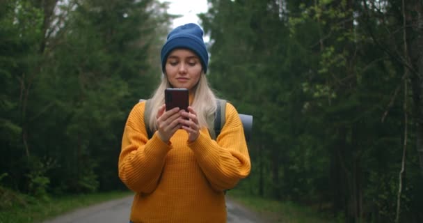 バックパック付きの黄色いセーターを着た女性旅行者が、美しい景色を眺めながら森の中の道を歩いています。森の中でアプリやインターネットを使って写真を撮る. — ストック動画