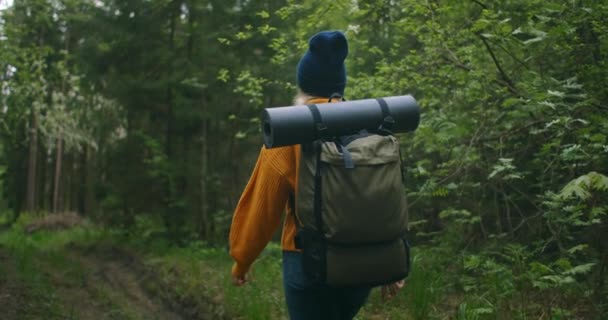 Kobieta podróżująca w żółtym swetrze z plecakiem idzie ścieżką w lesie, patrząc na piękne widoki. Kobieta podróżująca po lesie w swetrze z plecakiem — Wideo stockowe