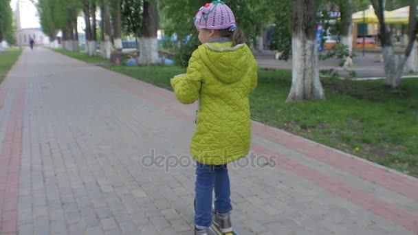 Niña con chaqueta verde montando una scooter. Disparando a Steadicam. Movimiento lento — Vídeo de stock