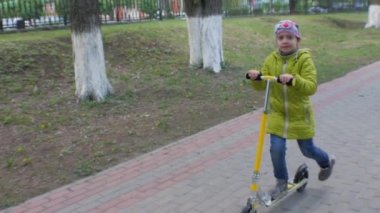 Yeşil ceket ve kot kaldırımda bir scooter sürme, küçük kız. Steadicam çekim. Ağır çekim.