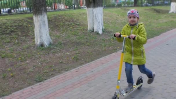 Malá holčička v zelené bundě a džíny jezdecké skútr na chodníku. Střelba Steadicam. Zpomalený pohyb. — Stock video