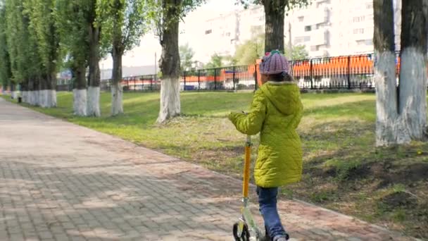 Klein meisje in de groene jas berijden van een scooter. Steadicam schieten. Slow motion — Stockvideo