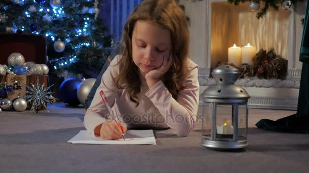 Une petite fille allongée par terre écrit une lettre au Père Noël. Cheminée de fond — Video