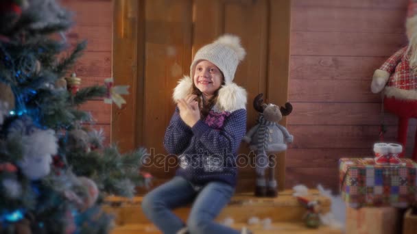 Όμορφο κοριτσάκι σε ζεστό καπέλο, φυσώντας τα χέρια του γύρω από το χριστουγεννιάτικο δέντρο — Αρχείο Βίντεο