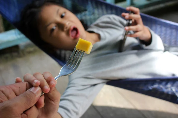 Dad feed a mango to his daughter who's only interested with the phone. — Stock Photo, Image