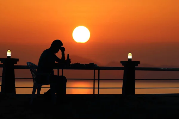 Silhouette sad man drinking beer at the beach with red sky sunse