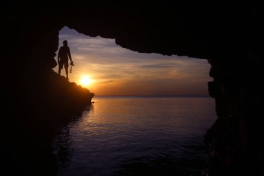 Fotoğrafçı silueti, alacakaranlık gökyüzü günbatımında deniz kenarındaki bir mağarada gün batımını yakalamak için kamerayı kullanıyor.