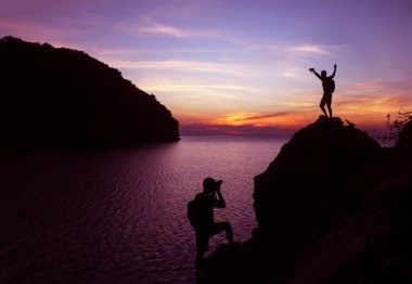 Fotoğrafçı silueti, deniz kenarındaki bir kayanın tepesinde kamera kullanıyor. Alacakaranlık gökyüzü günbatımı arka planı..