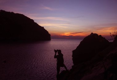 Fotoğrafçı silueti, deniz kenarındaki bir kayanın tepesinde kamera kullanıyor. Alacakaranlık gökyüzü günbatımı arka planı..