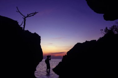 Fotoğrafçı silueti, alacakaranlık gökyüzü günbatımında deniz kenarındaki bir mağarada gün batımını yakalamak için kamerayı kullanıyor.