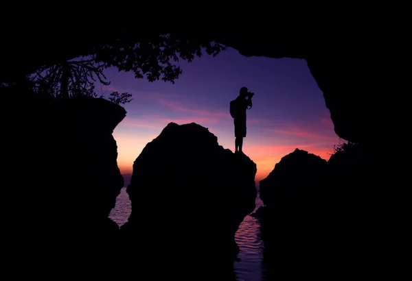 Man standing with a camera in front of a limestone cave by the sea at sunset, Photographer in front of the cave
