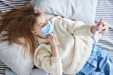 Selfie konsepti. Koruyucu tıbbi maskeli, kazaklı ve kot pantolonlu, yatakta uzanmış, akıllı telefondan kendi portresini yapan güzel bir kadın. Coronavirus covid-19 sırasında karantina ya da izolasyon.