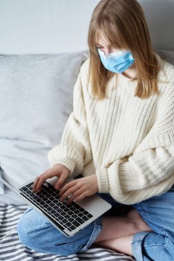 Medikal maskeli çekici bir kadın, evinde dizüstü bilgisayarla çalışıyor. Virüs hastalıkları sırasında karantinadaki defter ya da bilgisayar kullanarak çalışma, öğrenme, sörf yapma, sohbet etme kavramı.