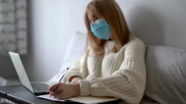 Beyaz kazaklı ve sağlık maskeli kız öğrenci dizüstü bilgisayarı ve defteriyle evde internet üzerinden ödev yapıyor. Coronavirus covid-19 karantina ve mesafe öğrenme kavramı