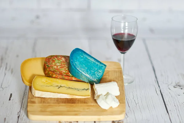 Copo de vinho tinto e variação de queijo em tábua de madeira — Fotografia de Stock