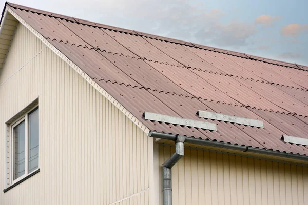 O telhado é de ardósia de telhado de amianto. As paredes da casa de tapume de plástico e telhado de ripas com chaminé. Folhas de coberturas onduladas e calha de chuva metálica . — Fotografia de Stock