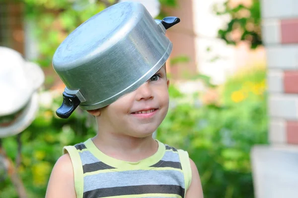 Un niño pequeño con una cacerola en la cabeza. niño con una cacerola. El niño feliz se complace. Un niño con un sombrero de cacerola . — Foto de Stock